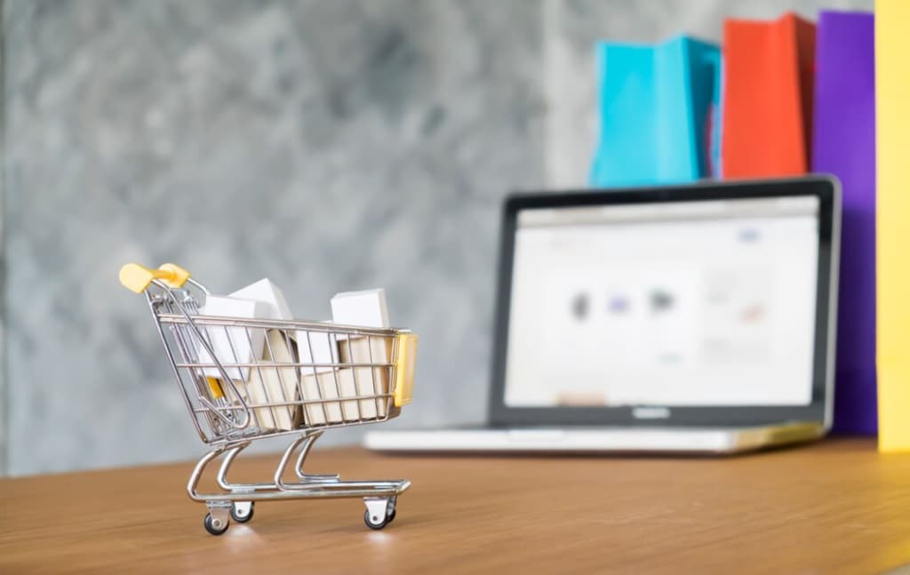 A mini shopping cart with packages in front of a laptop with shopping sites